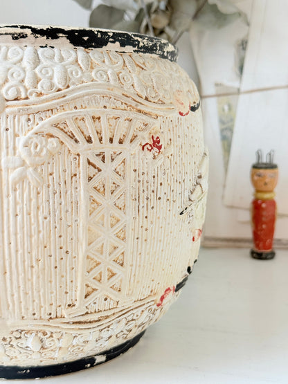 A close-up of a distressed cream-colored ceramic planter with carved architectural details and small red accents, shown on a white surface with a blurred doll in the background.