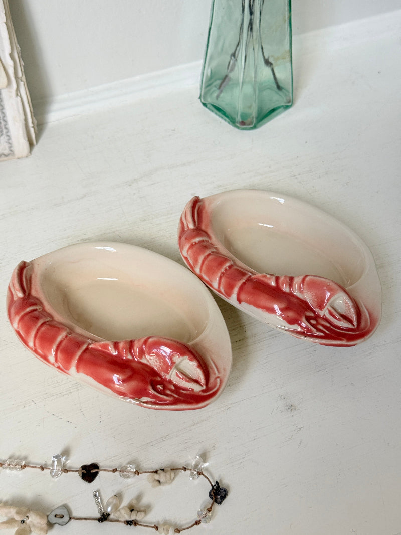 Two small oval ceramic dishes shaped like lobster tails, shown on a white tabletop with a blurred green glass vase in the background.