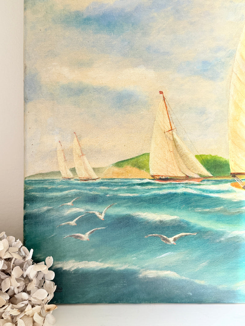 A square canvas painting of a sailing yacht race on a blue sea with flying seagulls and a green hill in the background, shown hanging on a white wall with dried flowers in the foreground.