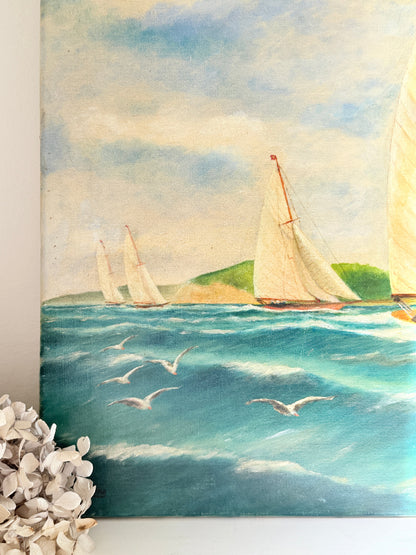 A square canvas painting of a sailing yacht race on a blue sea with flying seagulls and a green hill in the background, shown hanging on a white wall with dried flowers in the foreground.