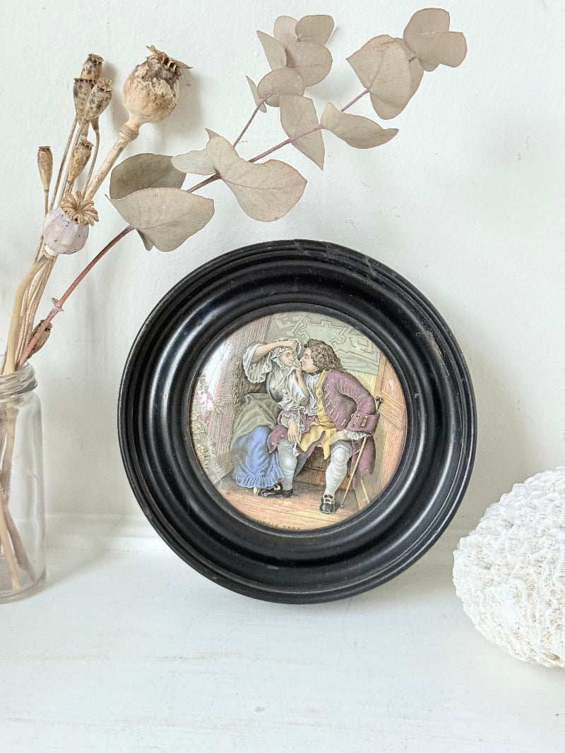 Antique Prattware pot lid in a black round frame with a detailed Victorian scene on a white surface.
