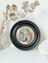 Antique Prattware pot lid in a black round frame with a detailed Victorian scene on a white surface.