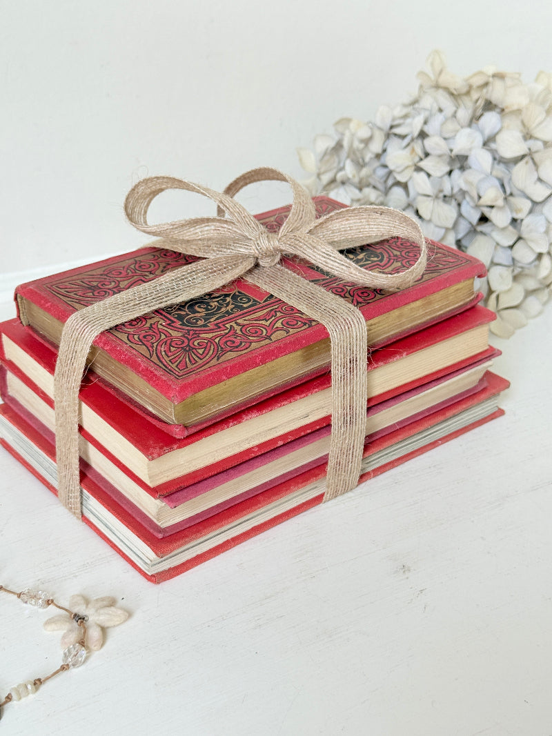 A stack of vintage-style red hardcover books tied with a beige jute ribbon bow, styled on a white surface with dried flowers in the background.