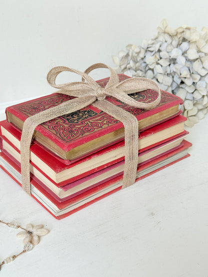 A stack of vintage-style red hardcover books tied with a beige jute ribbon bow, styled on a white surface with dried flowers in the background.