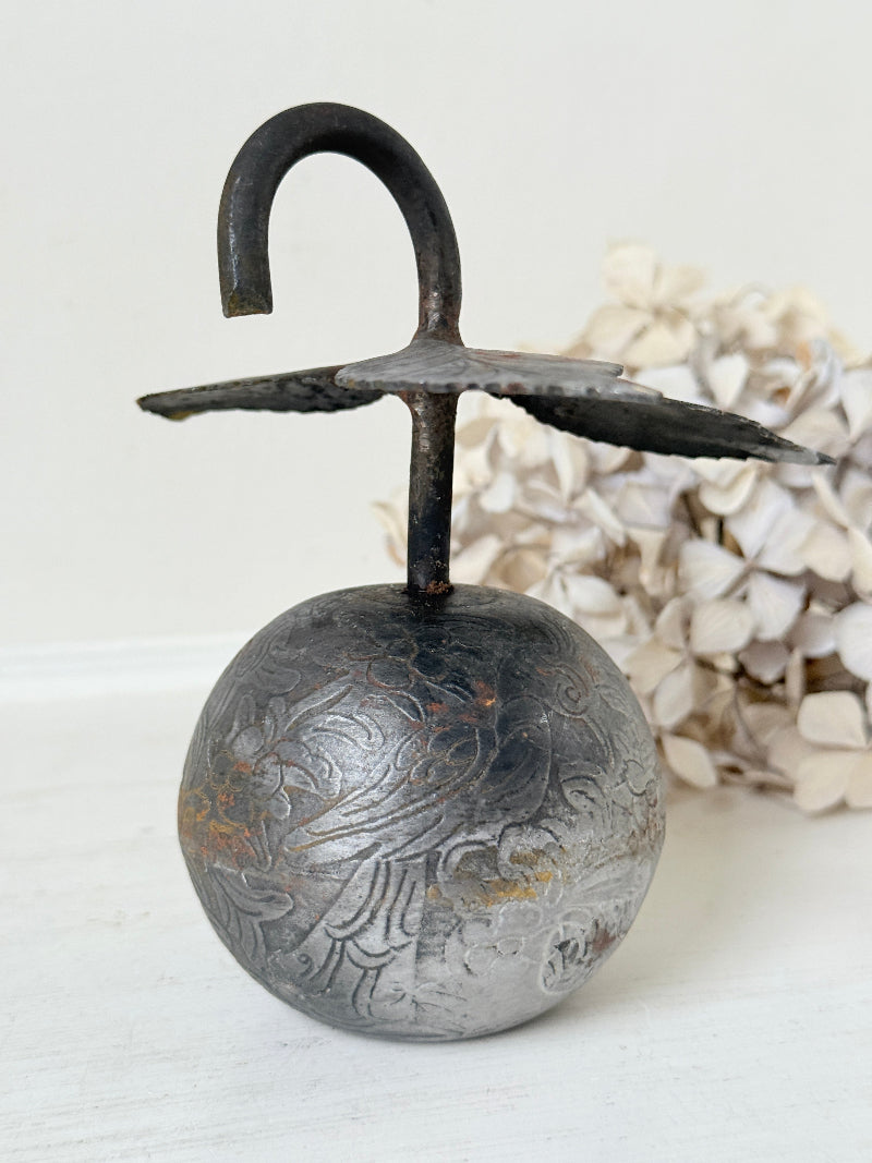 A vintage-style metal hanging ornament or decorative bell with an engraved silver-toned round body, dark handle, and wing-like top details, photographed against a soft white background with dried flowers behind it.
