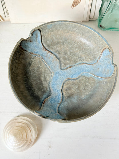 A handmade ceramic bowl with a rustic, hand-painted finish featuring a blue and gray river-like pattern, shown on a white wooden surface next to a small white seashell.