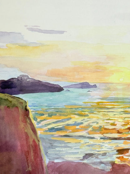 Framed coastal watercolour of sunset over sea with rocks in foreground, in gilt frame with pale mount, photographed on white surface.