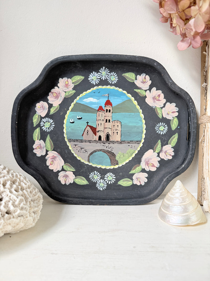 A black decorative serving tray with scalloped edges, featuring a hand-painted central scene of a castle on water surrounded by pink flowers and green leaves, styled on a white surface with seashells and dried flowers.