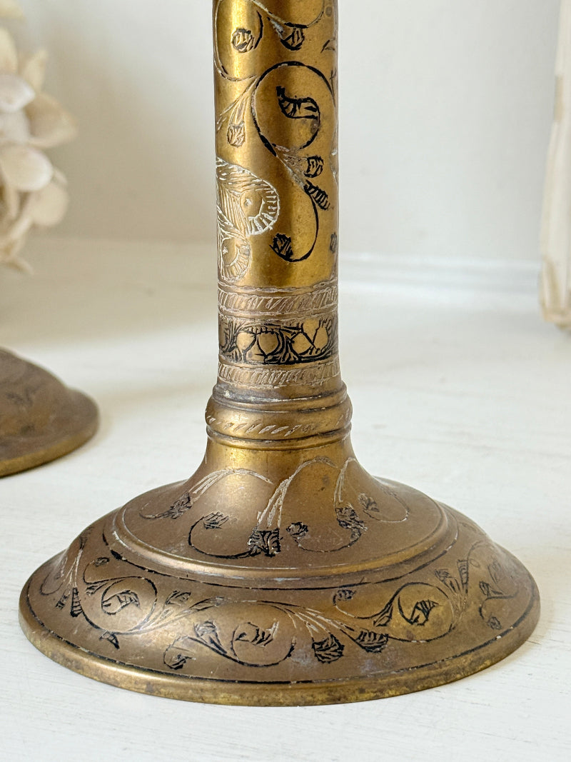 Pair of tall Indian engraved brass candlesticks in the Benares style with floral detailing and black taper candles on a white surface