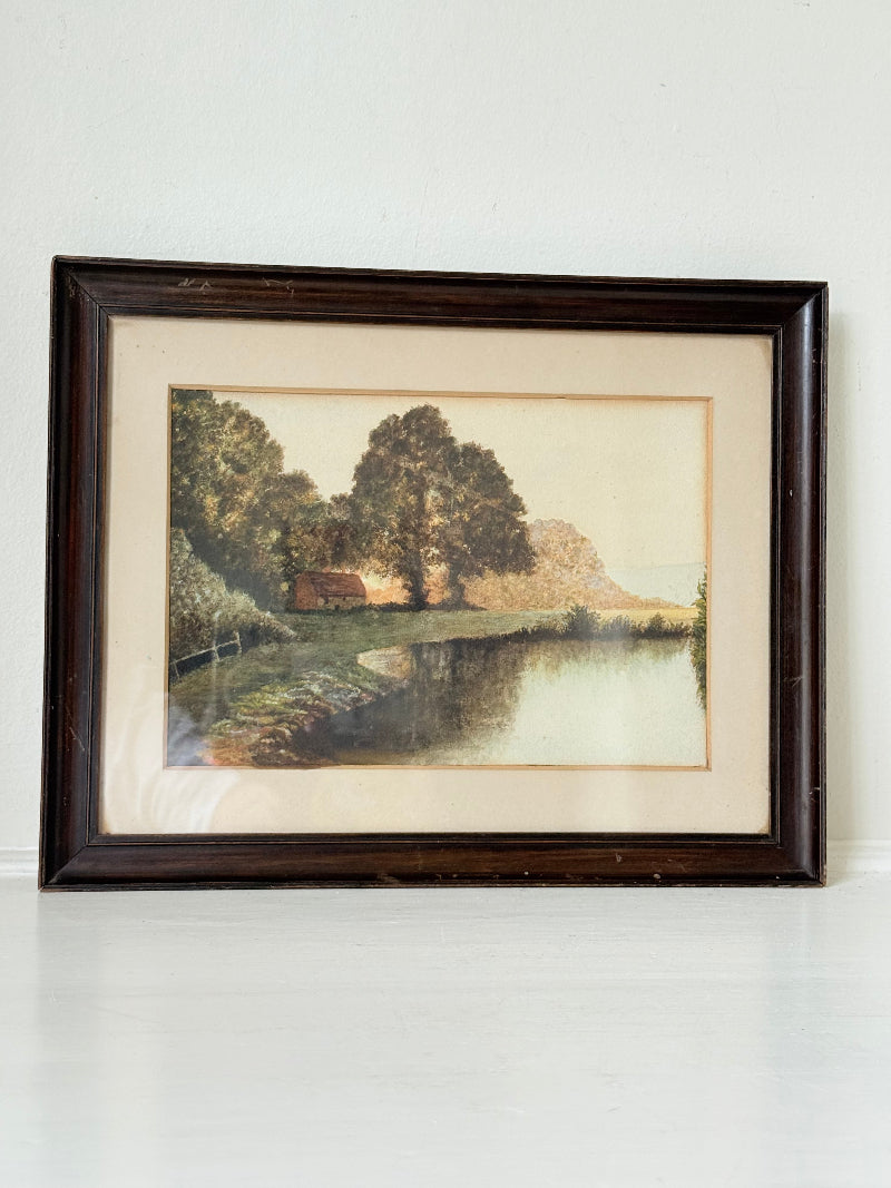Antique framed rural watercolour of river, trees, and red-roofed building, in dark wooden frame with cream mount, on white surface.