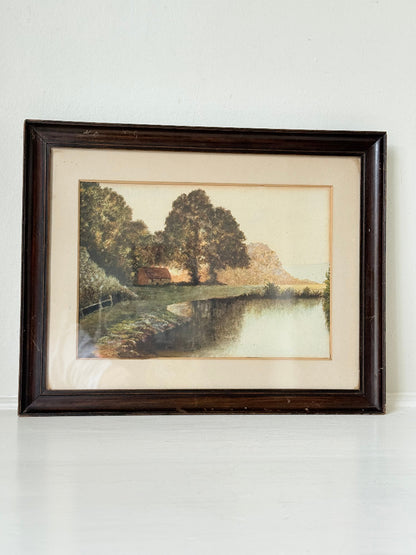 Antique framed rural watercolour of river, trees, and red-roofed building, in dark wooden frame with cream mount, on white surface.