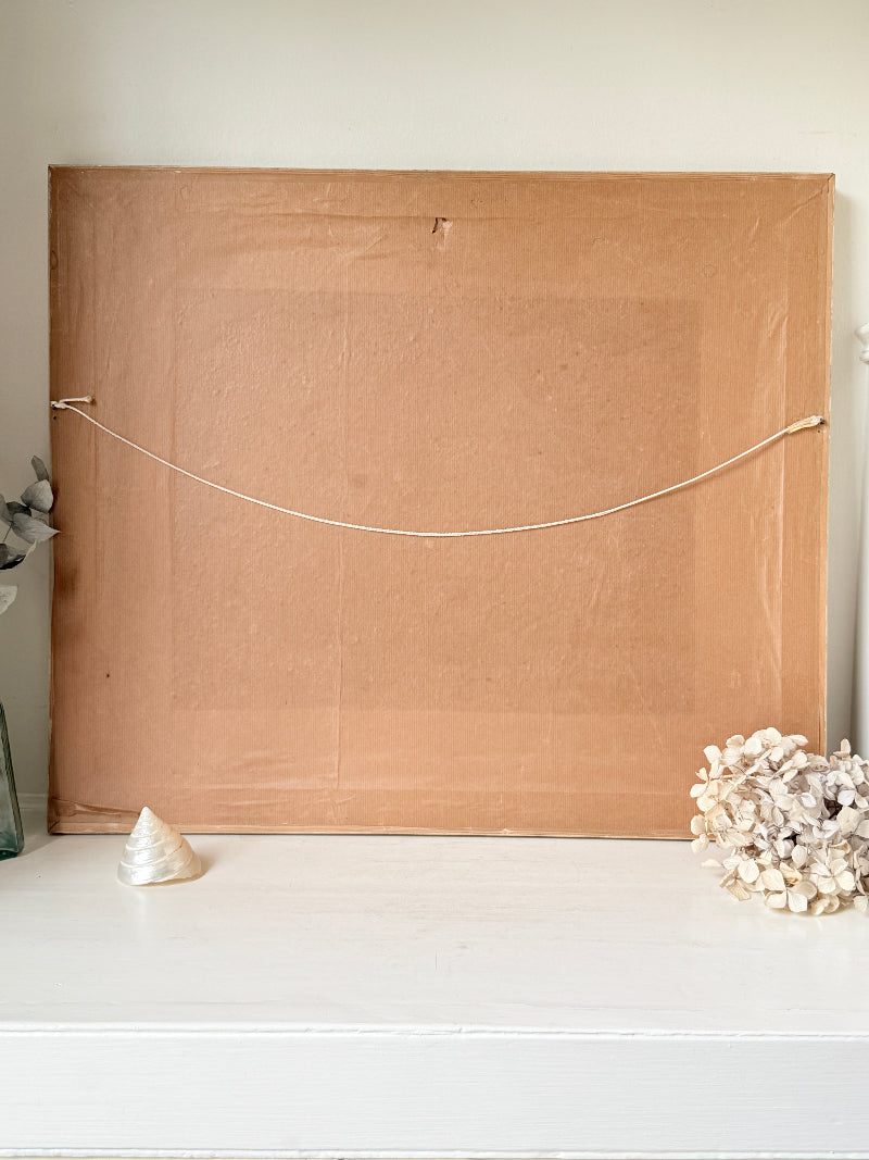 The back of a large square canvas or framed print, showing a tan cardboard backing with a white hanging cord stretched horizontally across it, displayed on a white shelf with decorative seashells.