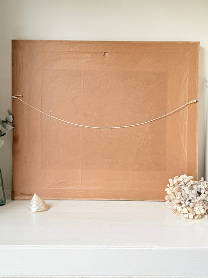 The back of a large square canvas or framed print, showing a tan cardboard backing with a white hanging cord stretched horizontally across it, displayed on a white shelf with decorative seashells.