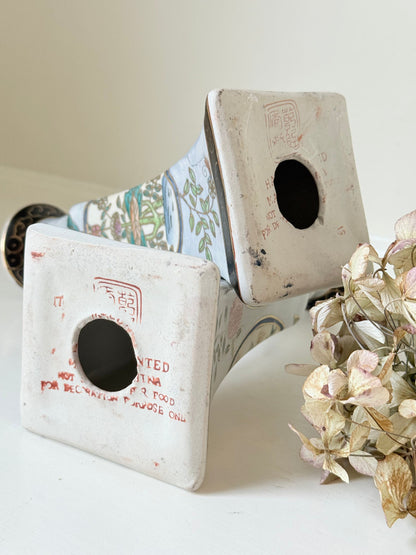 Pair of porcelain candle holders with hand-painted floral and vase motif in muted greens, pinks, and gold, styled with blush pink taper candles.