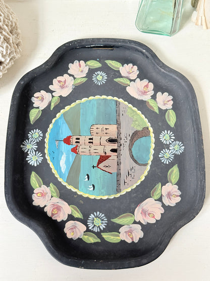 A square black decorative tray with scalloped edges, featuring a hand-painted central illustration of a castle by a blue lake, surrounded by pink flowers and green leaves on a white surface.