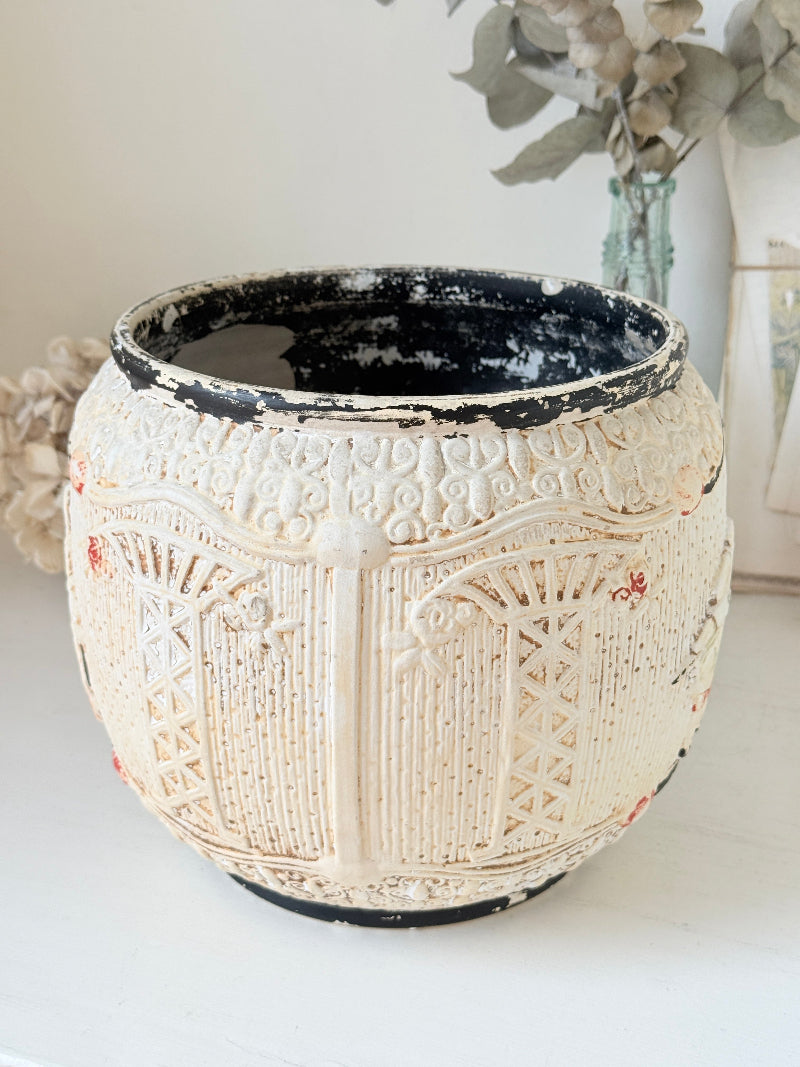 A round, vintage-style ceramic planter pot with a distressed cream-colored exterior featuring embossed architectural patterns and a black interior, set on a white surface with a soft-focus background.