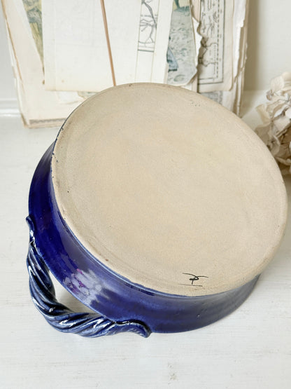 A handmade ceramic pot with a wide, unglazed beige clay top and a glossy cobalt blue glazed base and handle, resting on a white wooden surface.