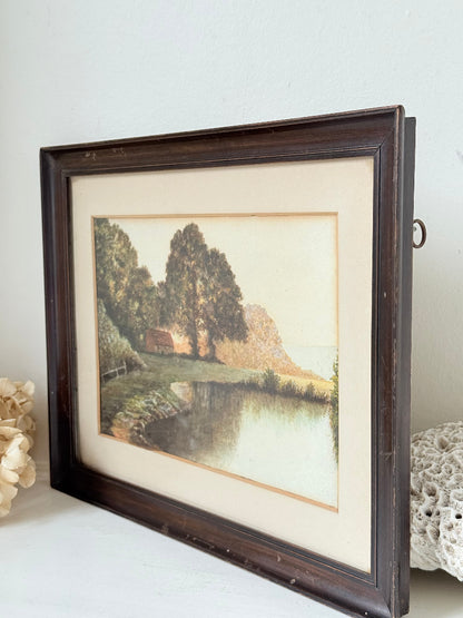 Antique framed rural watercolour of river, trees, and red-roofed building, in dark wooden frame with cream mount, on white surface.