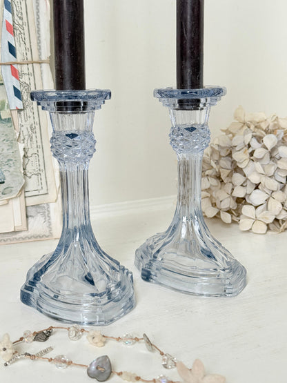 A pair of clear glass candlestick holders with a light blue tint and faceted detailing, shown holding black taper candles on a white surface with decorative props.
