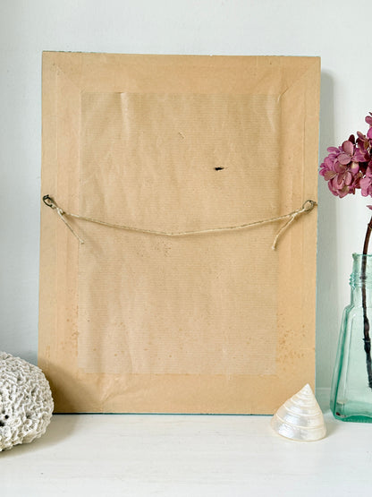 The back of a square tan canvas or wood wall art panel, secured with a rope and ring, displayed on a white surface with decorative seashells and a glass vase in the background.