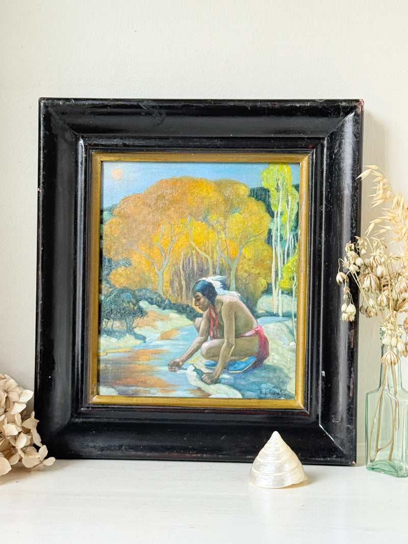 A framed vintage oil painting depicting a Native American man washing his face in a stream, surrounded by autumn trees. The painting is housed in a distressed black wooden frame with a gold inner border, displayed on a white surface with decorative dried flowers and a shell.