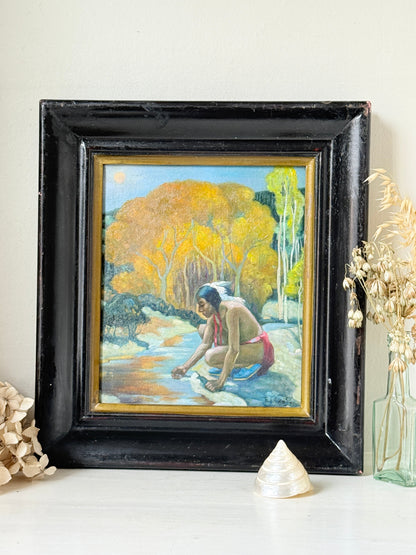 A framed vintage oil painting depicting a Native American man washing his face in a stream, surrounded by autumn trees. The painting is housed in a distressed black wooden frame with a gold inner border, displayed on a white surface with decorative dried flowers and a shell.