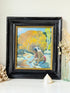A framed vintage oil painting depicting a Native American man washing his face in a stream, surrounded by autumn trees. The painting is housed in a distressed black wooden frame with a gold inner border, displayed on a white surface with decorative dried flowers and a shell.