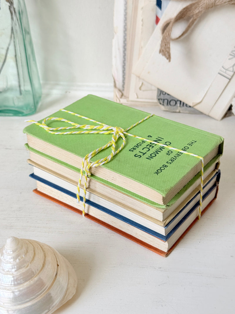 A stack of vintage hardcover books tied with yellow-and-white bakers twine, placed on a white wooden surface next to a seashell, with a glass bottle and other decorative items blurred in the background.