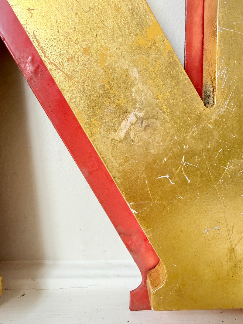 A close-up of a large decorative letter or panel with a distressed gold finish and red painted edges, photographed indoors against a light wall.