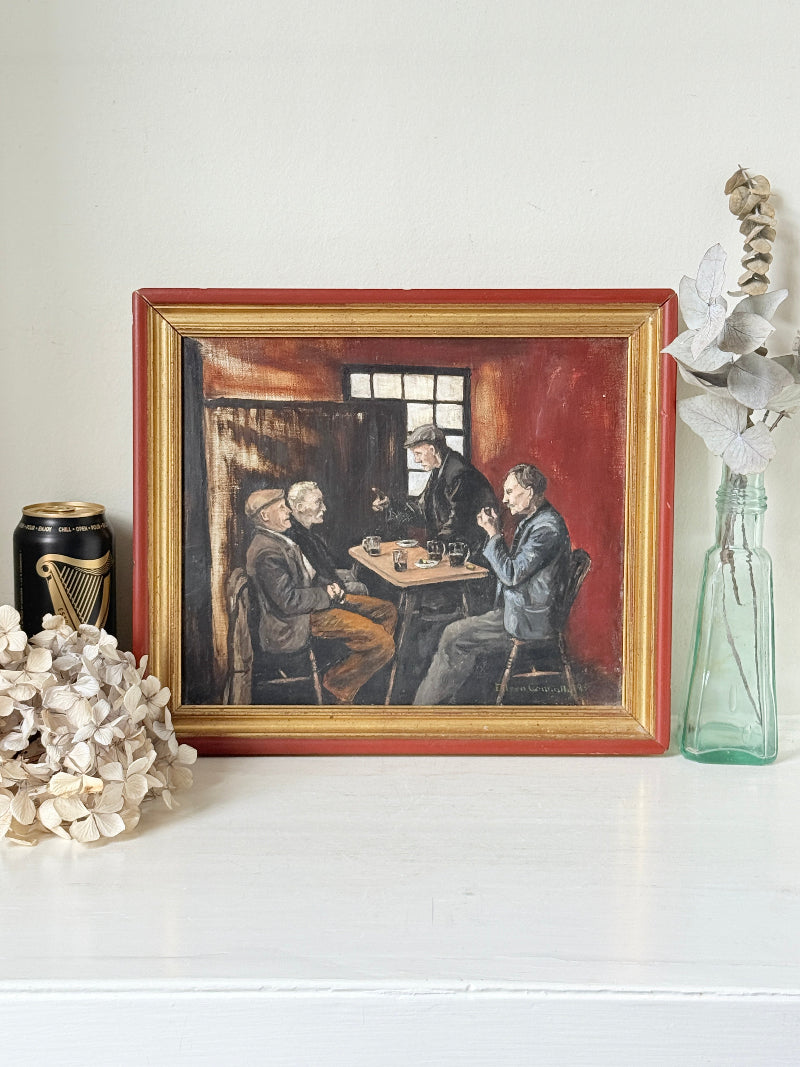 A framed vintage oil painting depicting three men playing chess in a dimly lit room, styled on a white shelf with decorative dried flowers and a glass bottle.