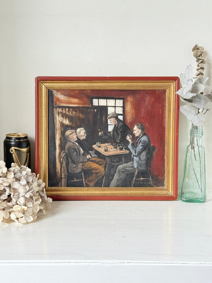 A framed vintage oil painting depicting three men playing chess in a dimly lit room, styled on a white shelf with decorative dried flowers and a glass bottle.