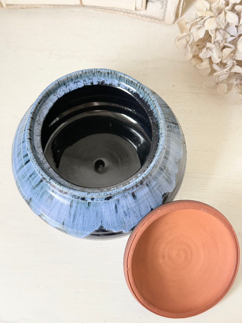 A ceramic jar with a blue and black glazed finish, shown open with its terracotta-colored lid resting beside it on a light-colored surface.