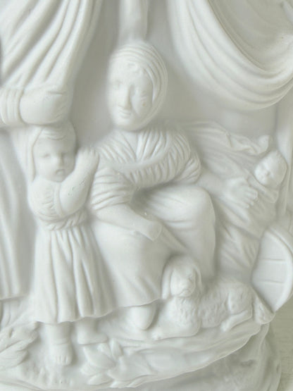 A close-up of a white sculptural relief featuring a seated woman, a standing child, and a baby, with draped fabric and soft carved details.
