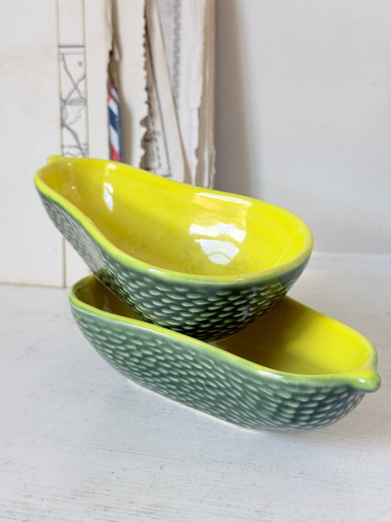 Two ceramic avocado-shaped bowls stacked on a white surface, with a textured green exterior and a bright yellow glazed interior.