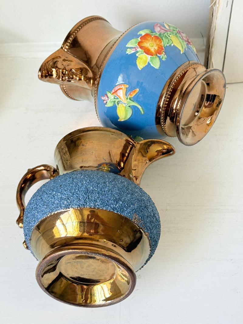 Two vintage-style decorative pitchers with metallic gold-toned bodies and handles, one featuring a blue floral glaze and the other a blue textured wrap, displayed on a white wooden surface.