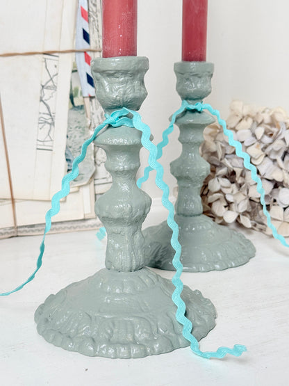 A pair of distressed sage green cast iron candlesticks holding red taper candles, decorated with a light blue wavy ribbon, set against a rustic background of framed prints and dried flowers.