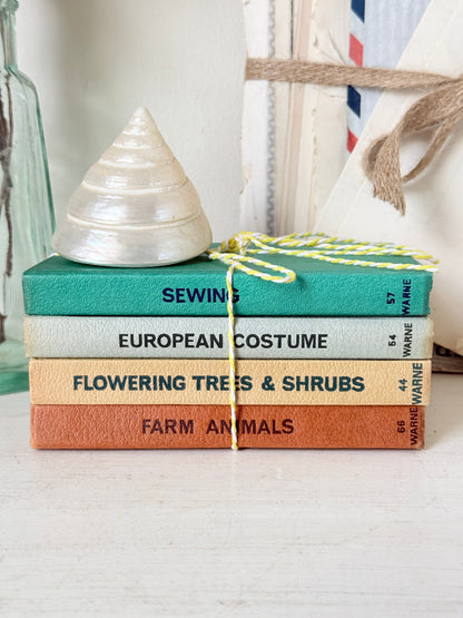 A stack of four vintage-style fabric books tied with yellow-and-white string, topped with a white seashell. The spines read Sewing, European Costume, Flowering Trees &amp; Shrubs, and Farm Animals.