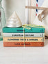 A stack of four vintage-style fabric books tied with yellow-and-white string, topped with a white seashell. The spines read Sewing, European Costume, Flowering Trees & Shrubs, and Farm Animals.
