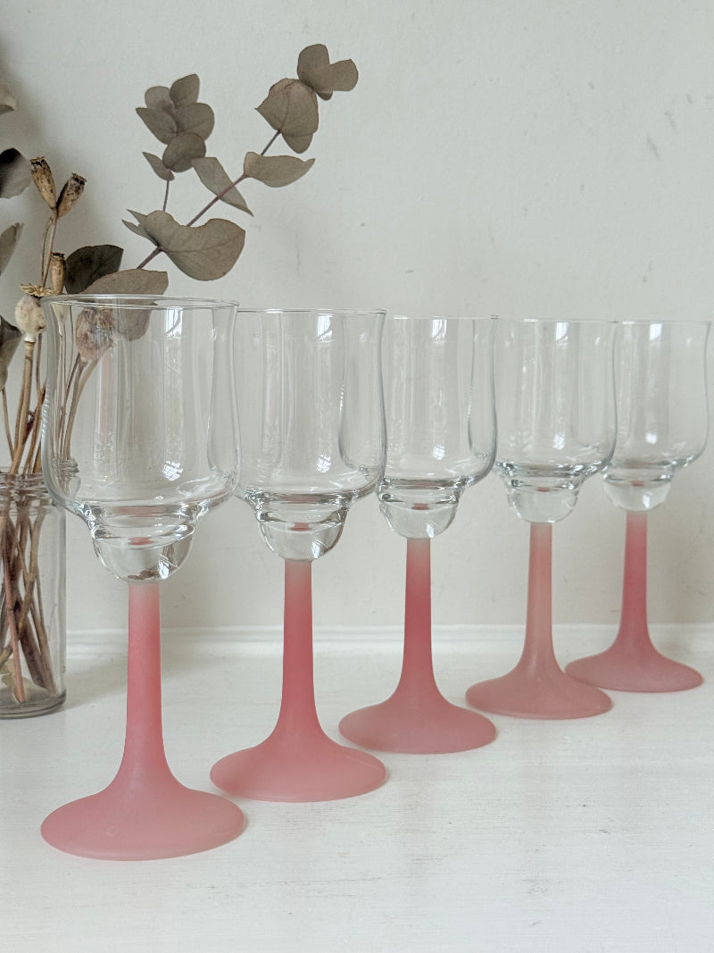 Set of five vintage wine glasses with clear bowls and pink stems photographed on a white surface.