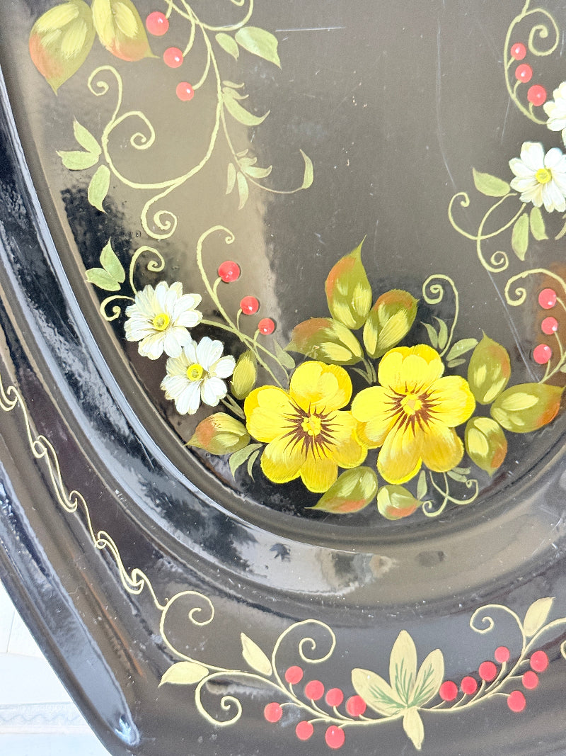 Close-up of a glossy black decorative tray or plate featuring hand-painted yellow flowers, white blossoms, green leaves, and red berries with gold vine scrollwork.