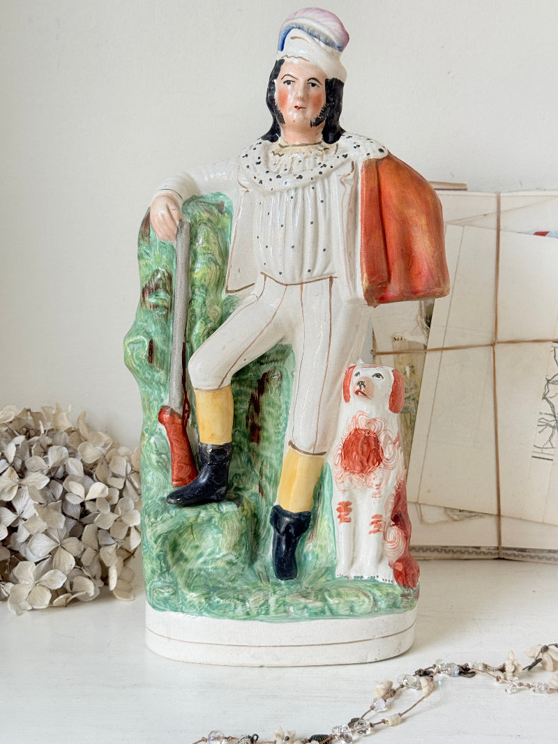A vintage ceramic figurine of a gentleman in riding attire, seated on a green tree stump with a dog at his side. The figure wears a white coat and breeches with yellow stockings and black boots, holding a sword and carrying an orange-brown satchel.