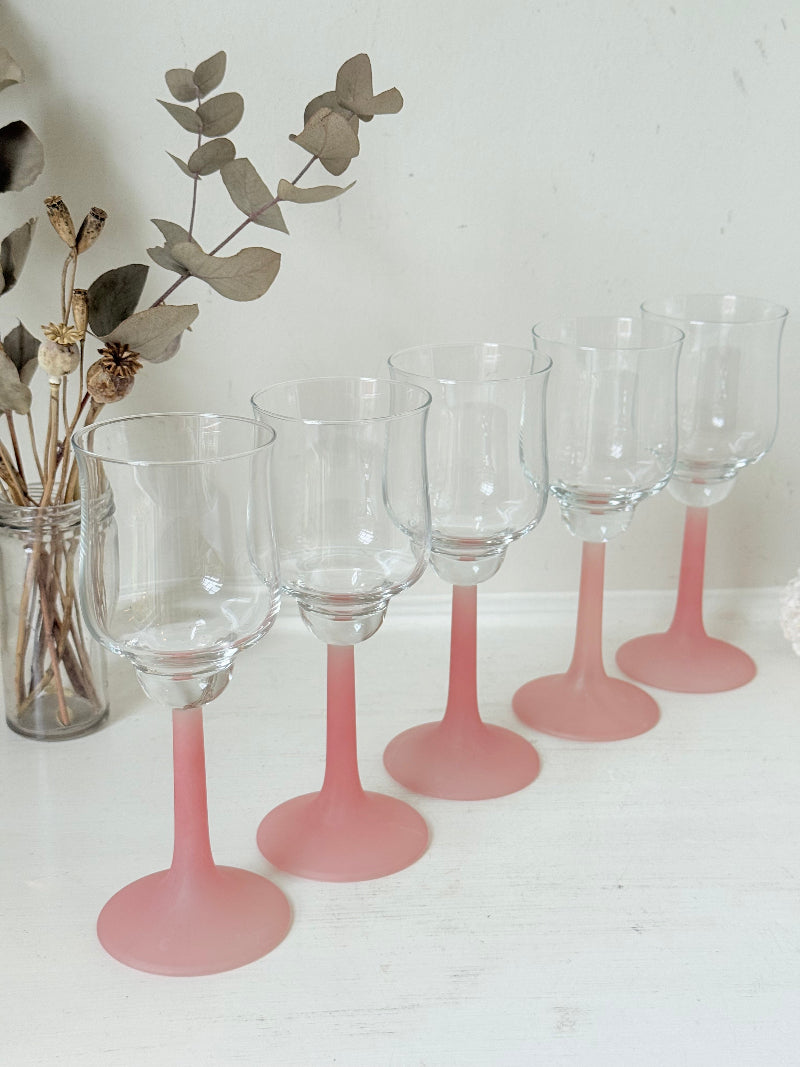 Set of five vintage wine glasses with clear bowls and pink stems photographed on a white surface.