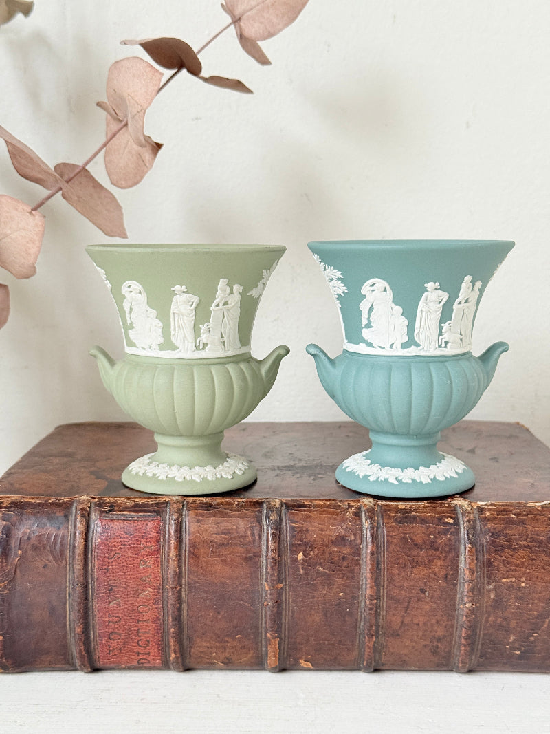 Pair of vintage Wedgwood Jasperware pedestal urns in sage green and pale blue, decorated with white neoclassical figures, styled on an antique leather book.