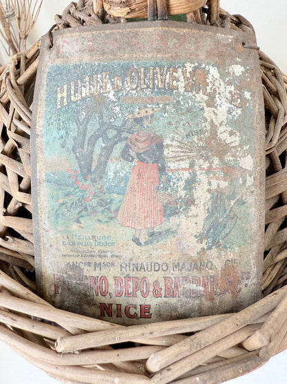 antique French demi john olive oil jar in wicker basket with green glass bottle and painted metal label, displayed on white surface