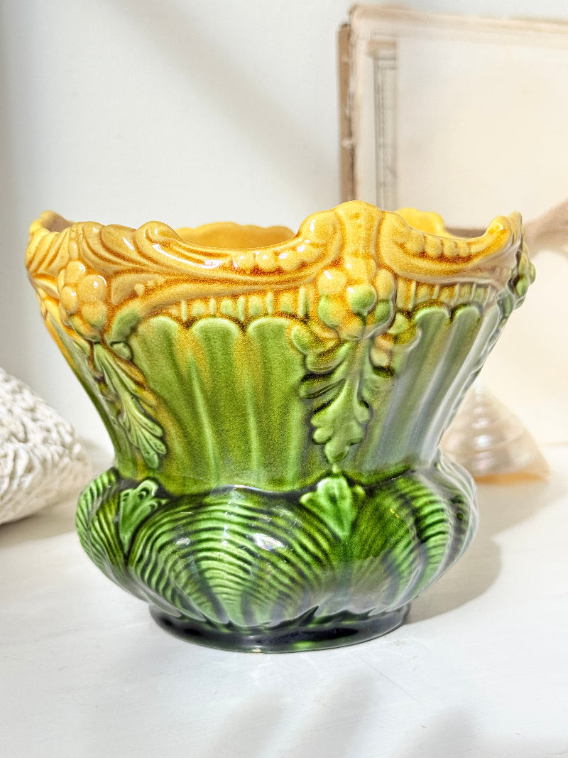 An ornate ceramic planter pot with a sculpted leaf design, featuring a glossy yellow and green glaze on a light surface.