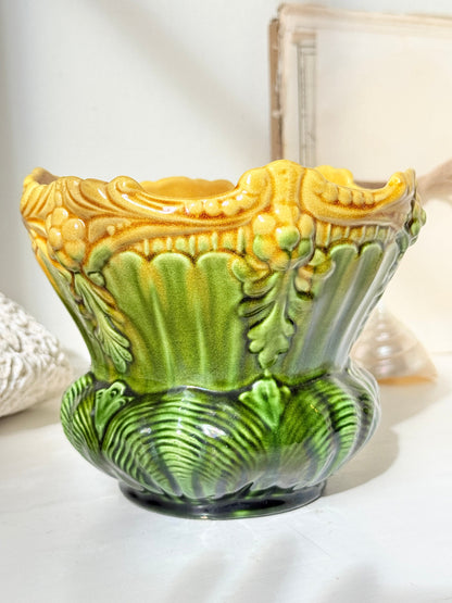 An ornate ceramic planter pot with a sculpted leaf design, featuring a glossy yellow and green glaze on a light surface.