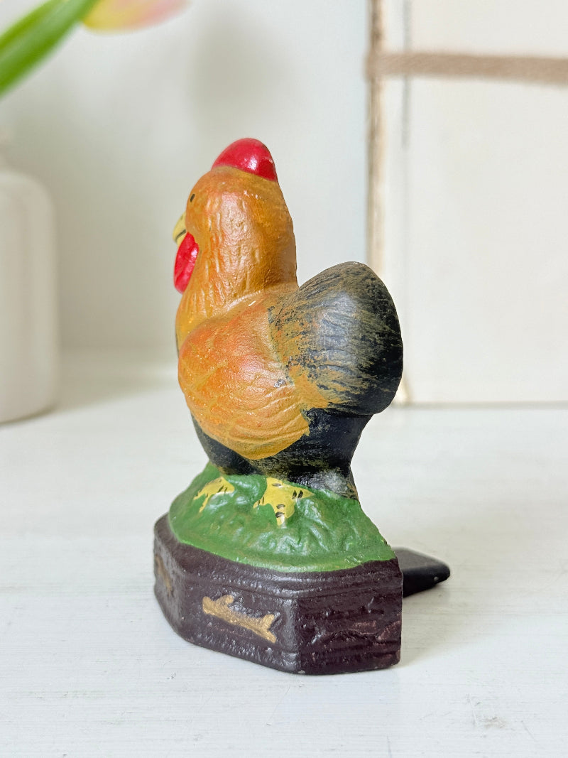 A vintage hand-painted wooden rooster figurine standing on a small green hill atop a dark brown base, photographed on a white tabletop with a softly blurred indoor background.