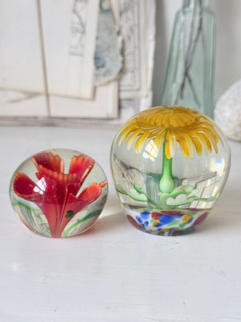 Pair of yellow and red flower glass paperweights with green stems, photographed on a white surface