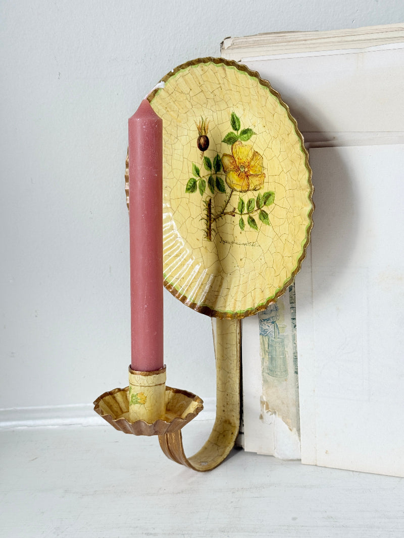 Vintage wall-mounted candle holder with a scalloped yellow ceramic panel featuring a yellow flower and green leaves, holding a pink taper candle, set against a white wall and shelf.