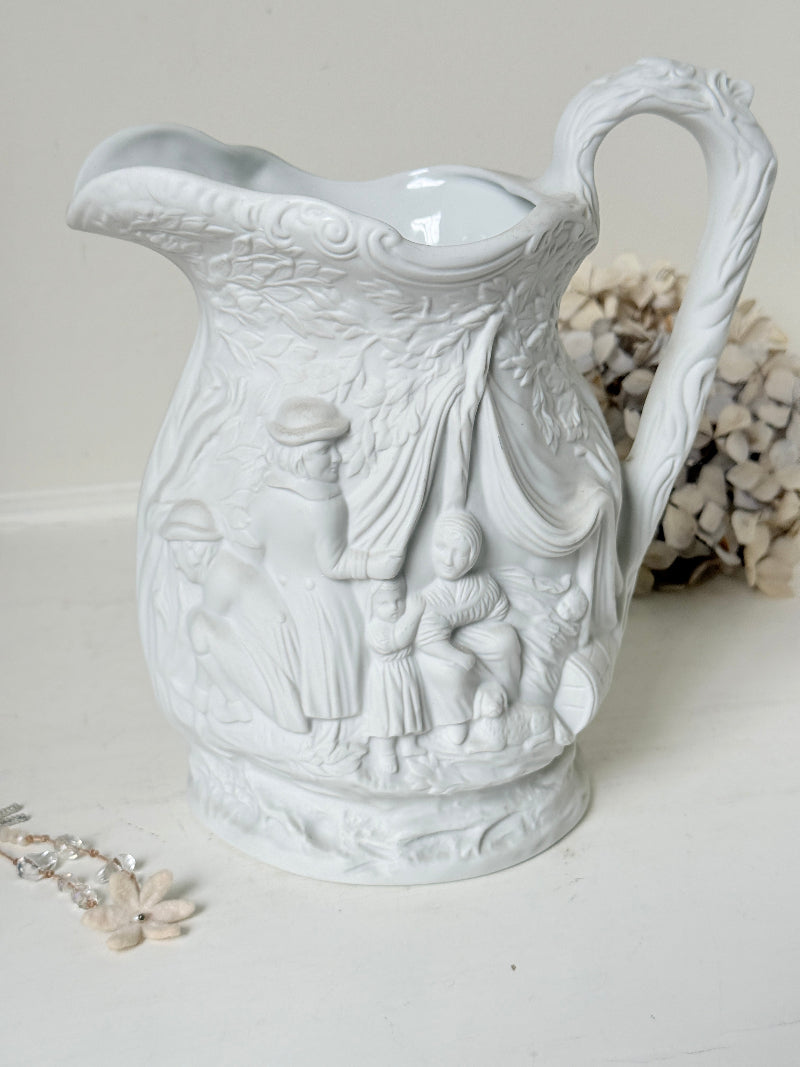 A white ceramic pitcher with embossed classical figures and draped details, set against a neutral background with dried flowers and a pearl necklace prop.